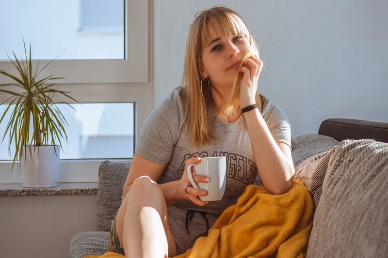 Young woman on a sofa with a mustard throw over her legs, mug in hand, in the sunlight by a window