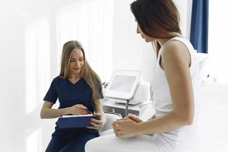 A young woman in navy medical scrubs, holding a folder and a pen, talking with a patient in a white vest top sitting opposite her in a bright treatment room