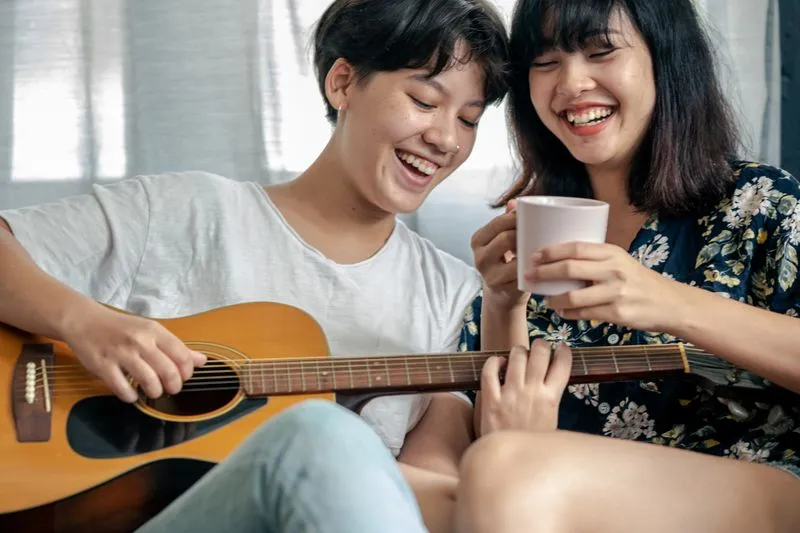 Two young women laughing together, one playing guitar while the other holds a cup of coffee