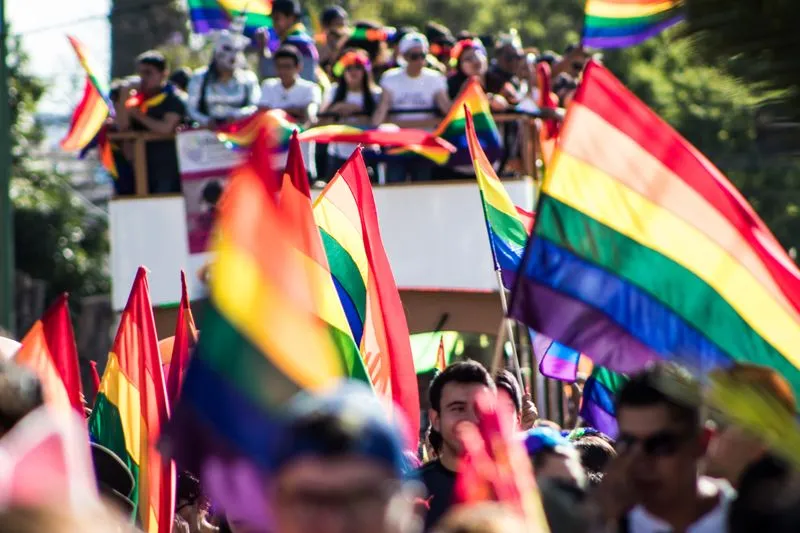 A large rainbow flag held aloft above a crowd at a sunny Pride march