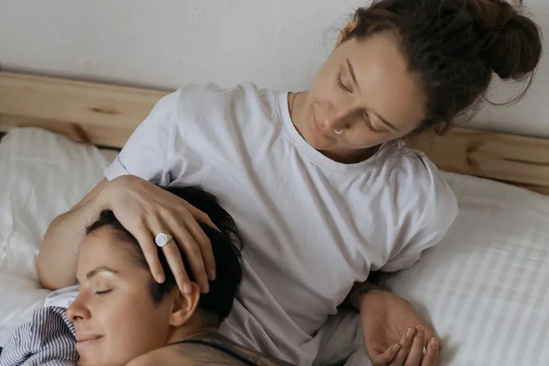 Two women in bed with pale sheets, one lying with her eyes closed while the other gently strokes her hair