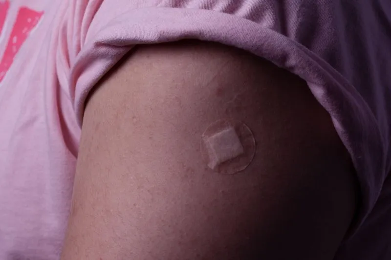 A small square dressing at the centre of a round adhesive patch, on the upper arm of a young woman after an injection