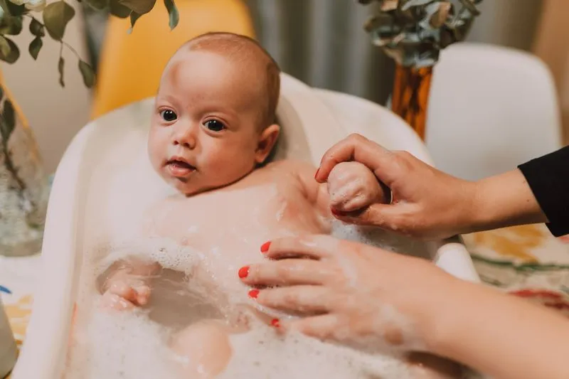Baby sitting in a small tub of lukewarm water, held by an adult's hand