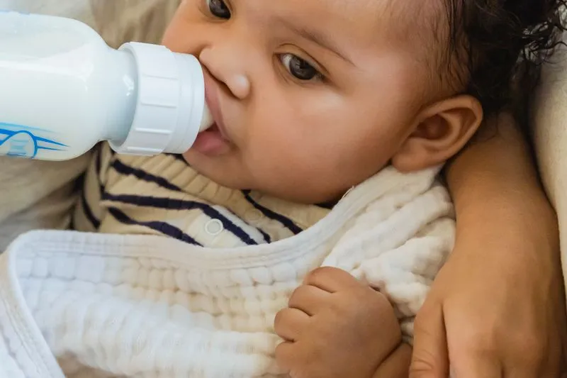 Baby drinking from a bottle, cradled in an adult's arm indoors