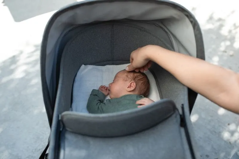 Baby asleep in a pram's carrycot with the hood up, an adult's hand resting on their forehead