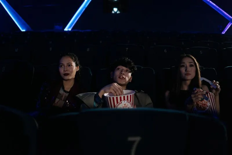 Three friends sitting in a cinema, two of them holding tubs of popcorn, their faces lit by the screen
