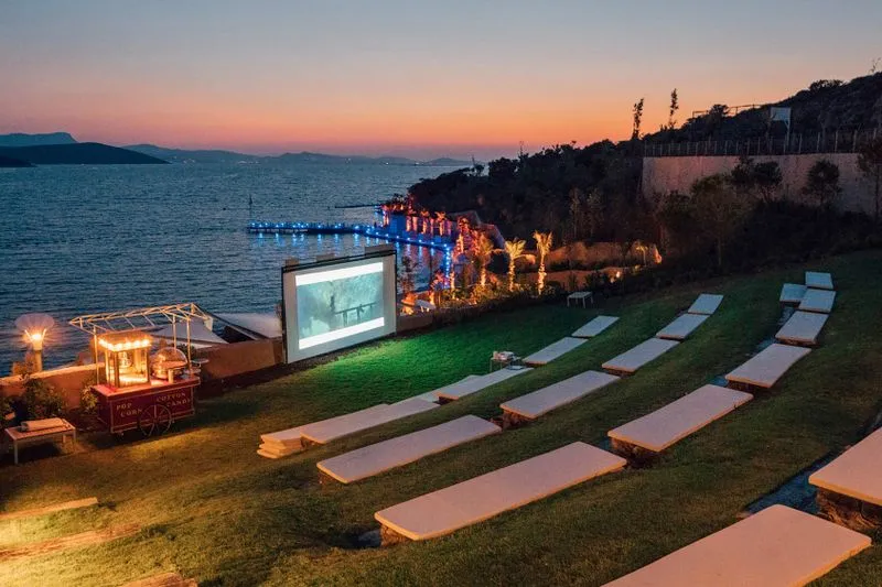 Rows of loungers facing a large outdoor cinema screen by the water at dusk