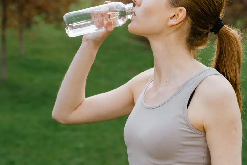 A female athlete in a sports bra and tank top drinking from her water bottle in a park