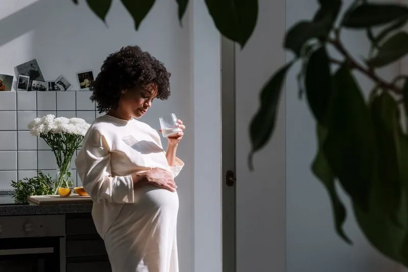 A pregnant woman holding a glass, one hand resting on her bump, in a bright kitchen next to a vase of white flowers