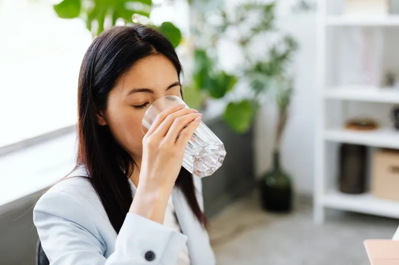 A young woman drinking a glass of cold water by a sunny window