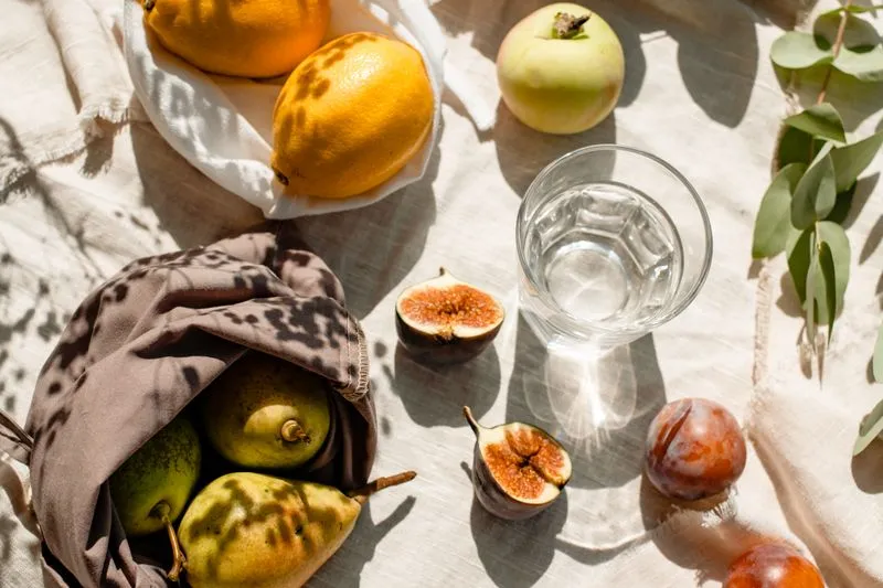 Glass of water surrounded by summer fruit — figs, lemons, an apple, plums — on a picnic cloth