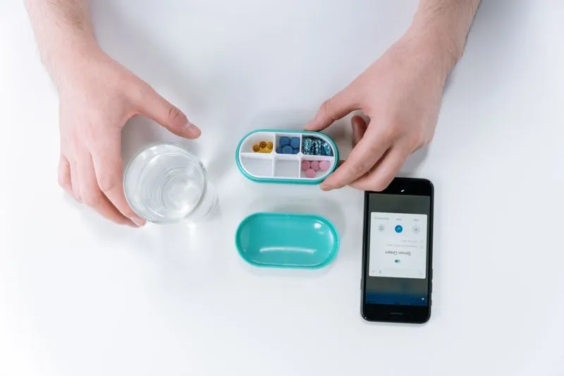 Small pill box held open in one hand, next to a glass of water and a smartphone