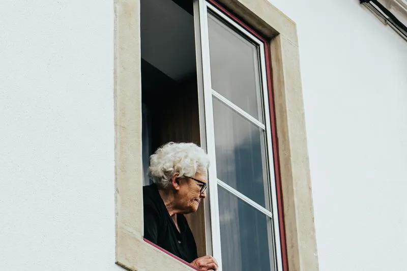 Older woman with white hair and glasses leaning out of an open window in a white facade
