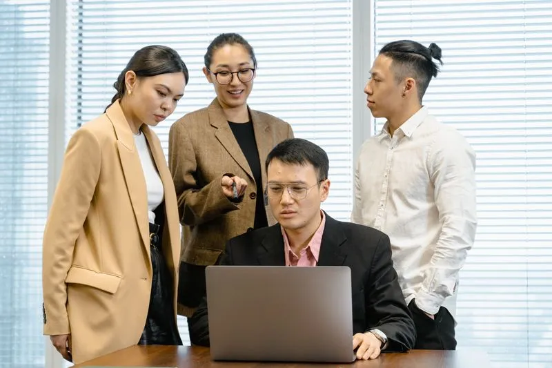 Four colleagues gathered around a laptop in an office, one woman in a blazer explaining something with an animated gesture