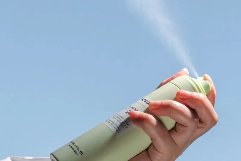 Hand holding a pale green spray deodorant can, misting a fine spray against a blue sky