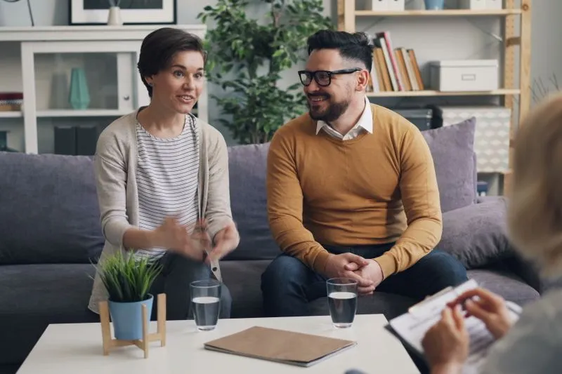 Couple sitting side by side facing a counsellor in a bright office, during a session