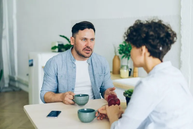 Young couple sitting at a kitchen table, facing each other in conversation over two mugs