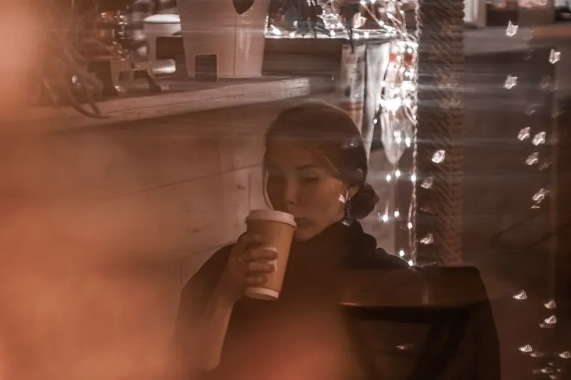 Woman sitting alone in a café, sipping from a takeaway cup, seen through the window