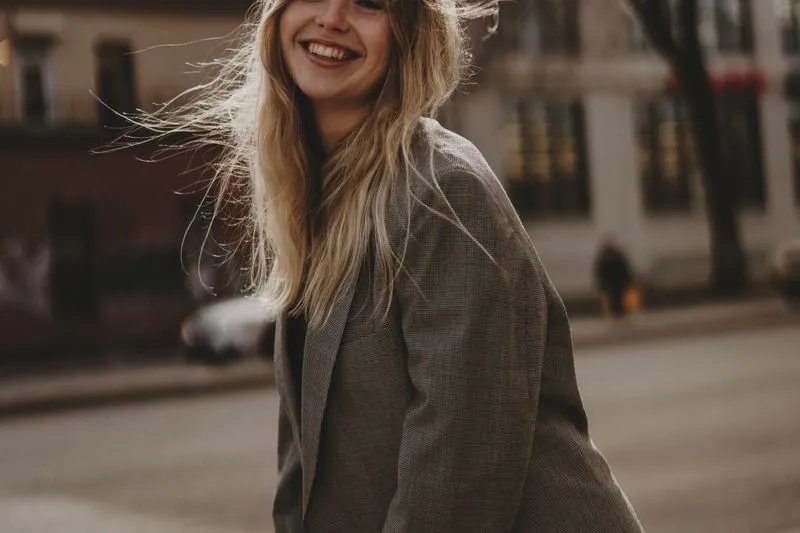 Woman laughing alone on a city street, hair in the wind, wearing a check blazer