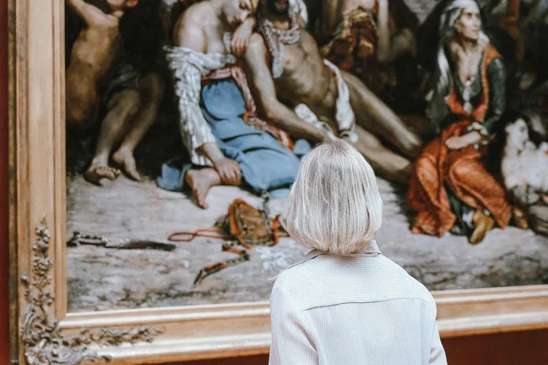 Woman with short platinum-blonde hair, seen from behind, standing alone before a large old painting in a gilded frame