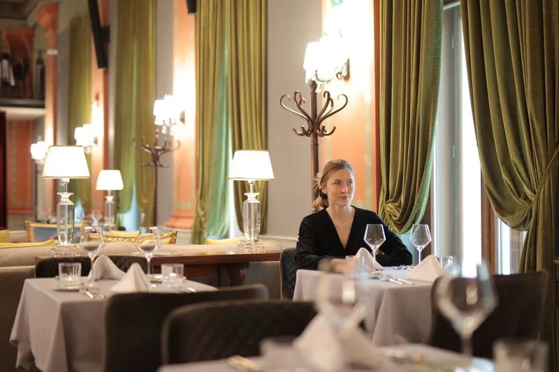 Woman sitting alone at a table in an elegant restaurant with green velvet curtains, looking relaxed