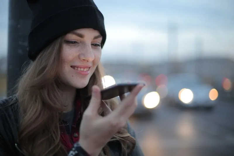 Young woman in a black beanie recording a voice message on her phone at the roadside at dusk, car headlights blurred behind her