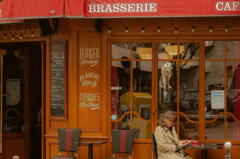 Woman in a beige trench coat and glasses sitting alone outside a brasserie, a red cup in front of her and her phone in her hand